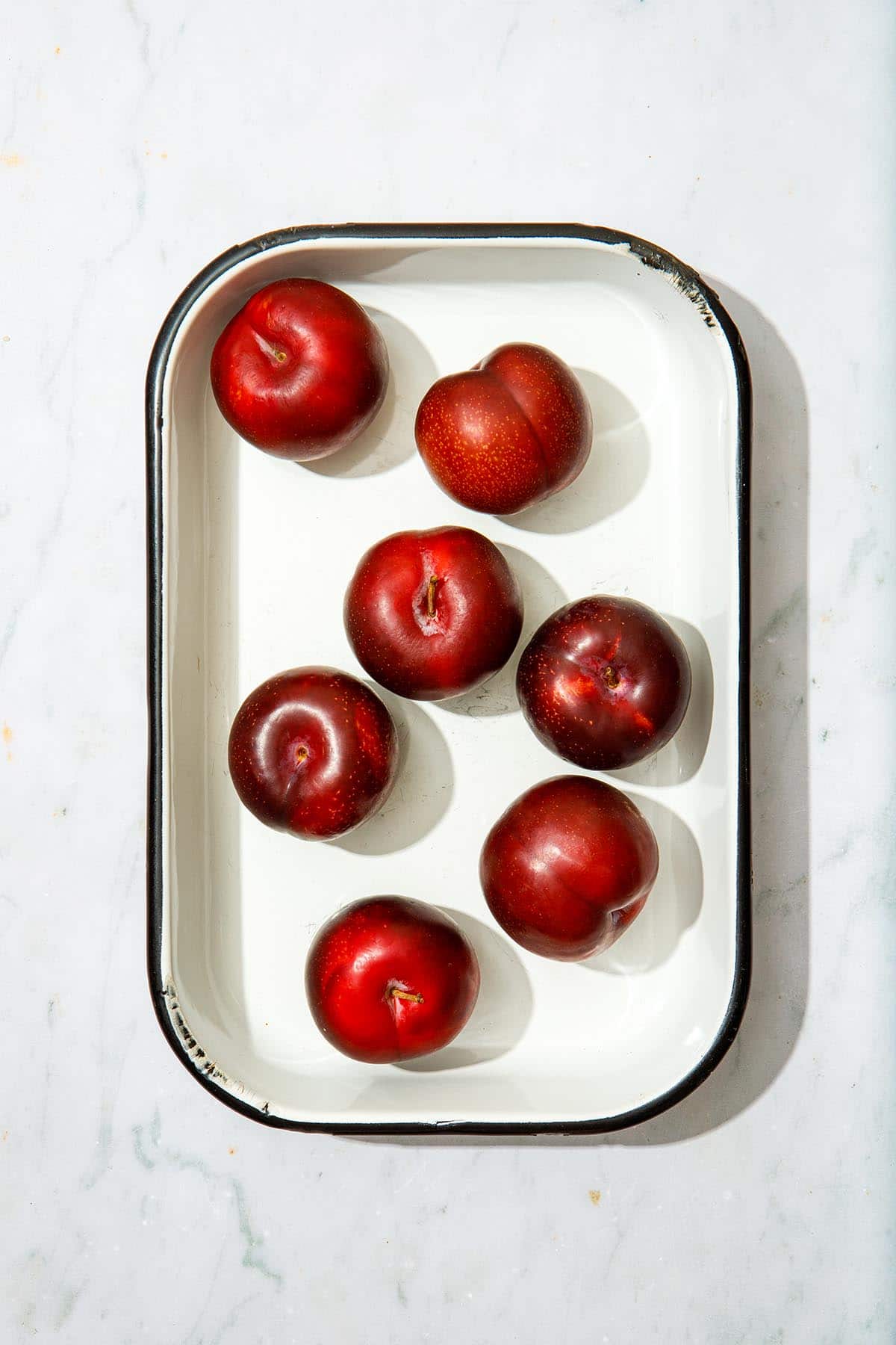 Seven fresh plums inside a small white rectangular enamel high-sided tray with black trim around the edges.