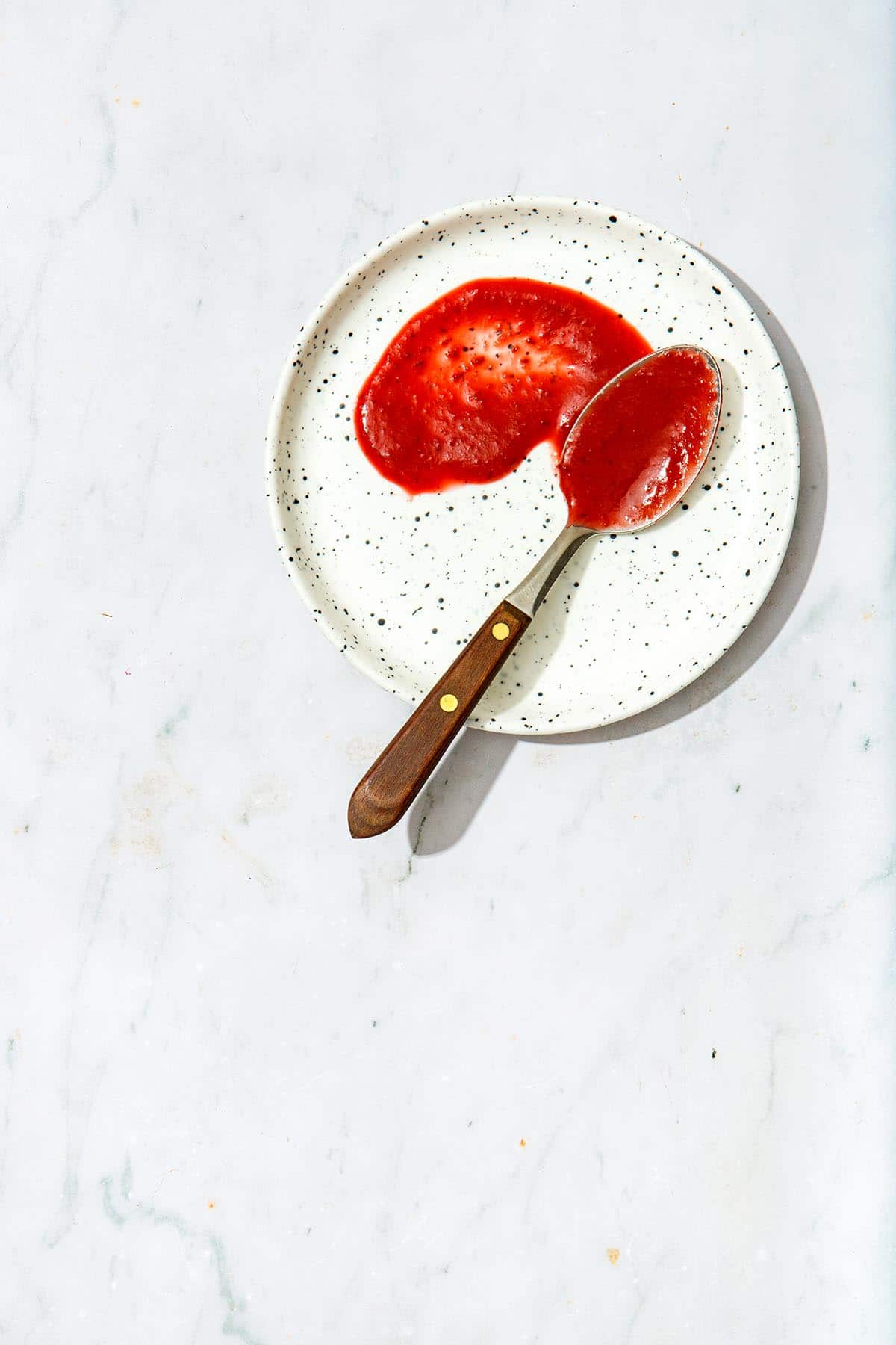 A spoonful of vegan BBQ sauce smeared artfully on a small white speckled plate on a marble background.