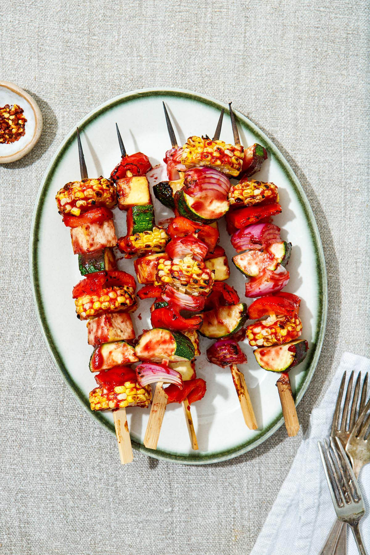 Grilled veggie skewers, brushed with vegan BBQ sauce, laying on a white oval platter with green trim.