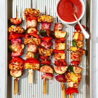Grilled veggie skewers on a metal tray alongside a small bowl of vegan BBQ sauce.
