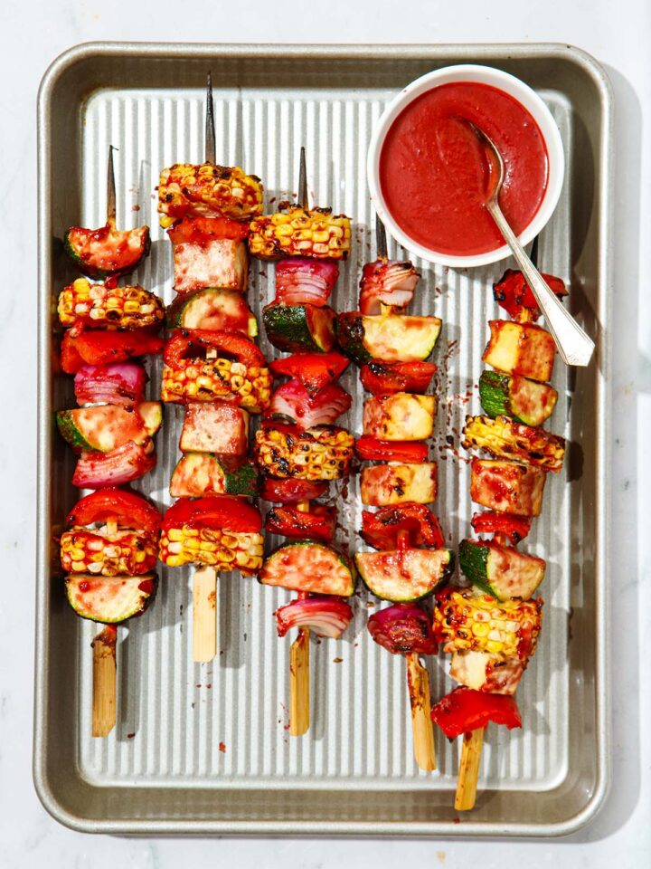 Grilled veggie skewers on a metal tray alongside a small bowl of vegan BBQ sauce.