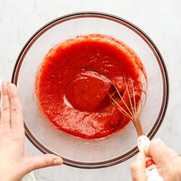 A hand using a whisk to mix vegan BBQ sauce in a large glass bowl.