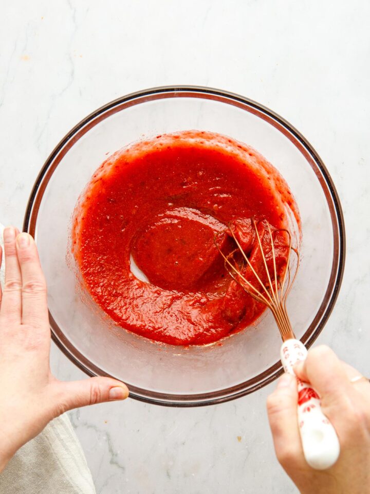 A hand using a whisk to mix vegan BBQ sauce in a large glass bowl.