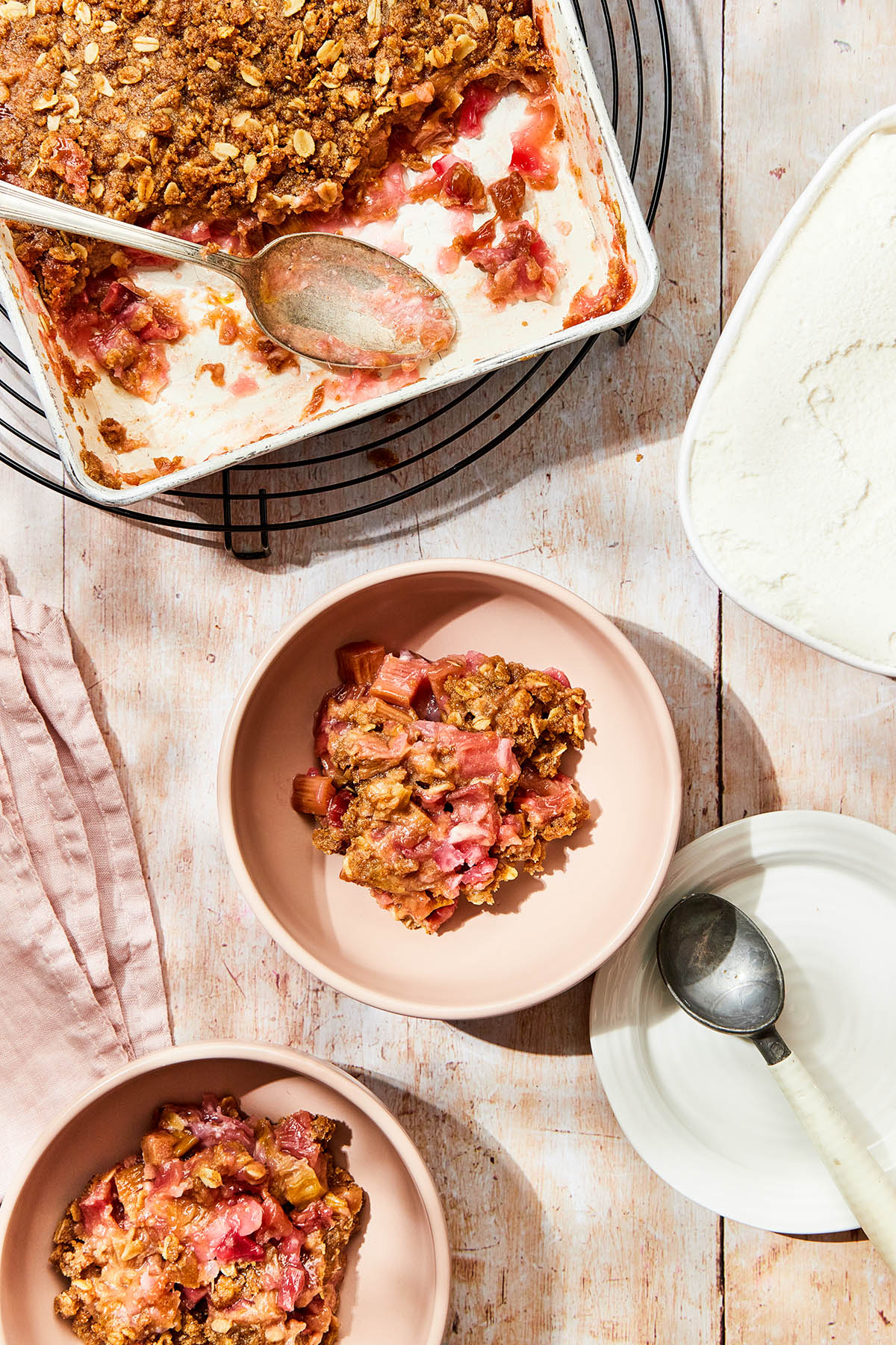 A pan of gluten-free rhubarb crisp on a wood table along with two bowl of crisp, a carton of un-scooped ice cream and an ice cream scoop.