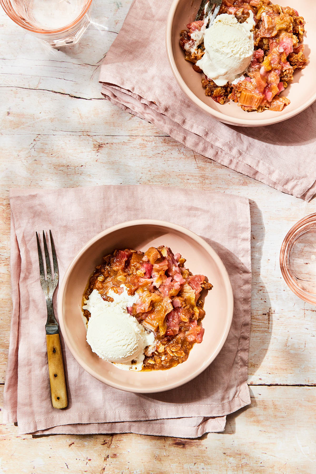 Two bowls of gluten-free rhubarb crisp topped with ice cream on a wood table.