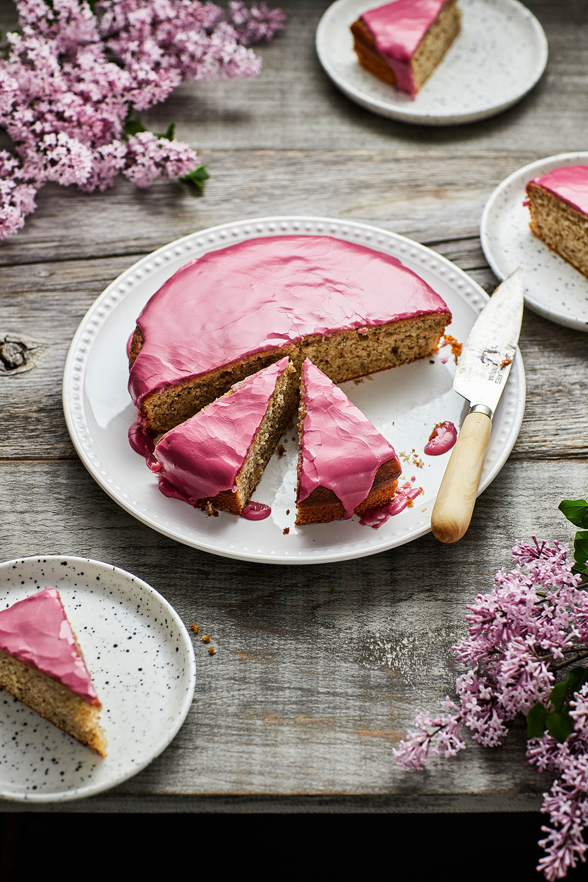 A sliced Earl Grey yogurt cake with pinkish-purple blackberry glaze on a grey weathered table. There are slices of cake on small playtes and lilacs on the table around the cake.
