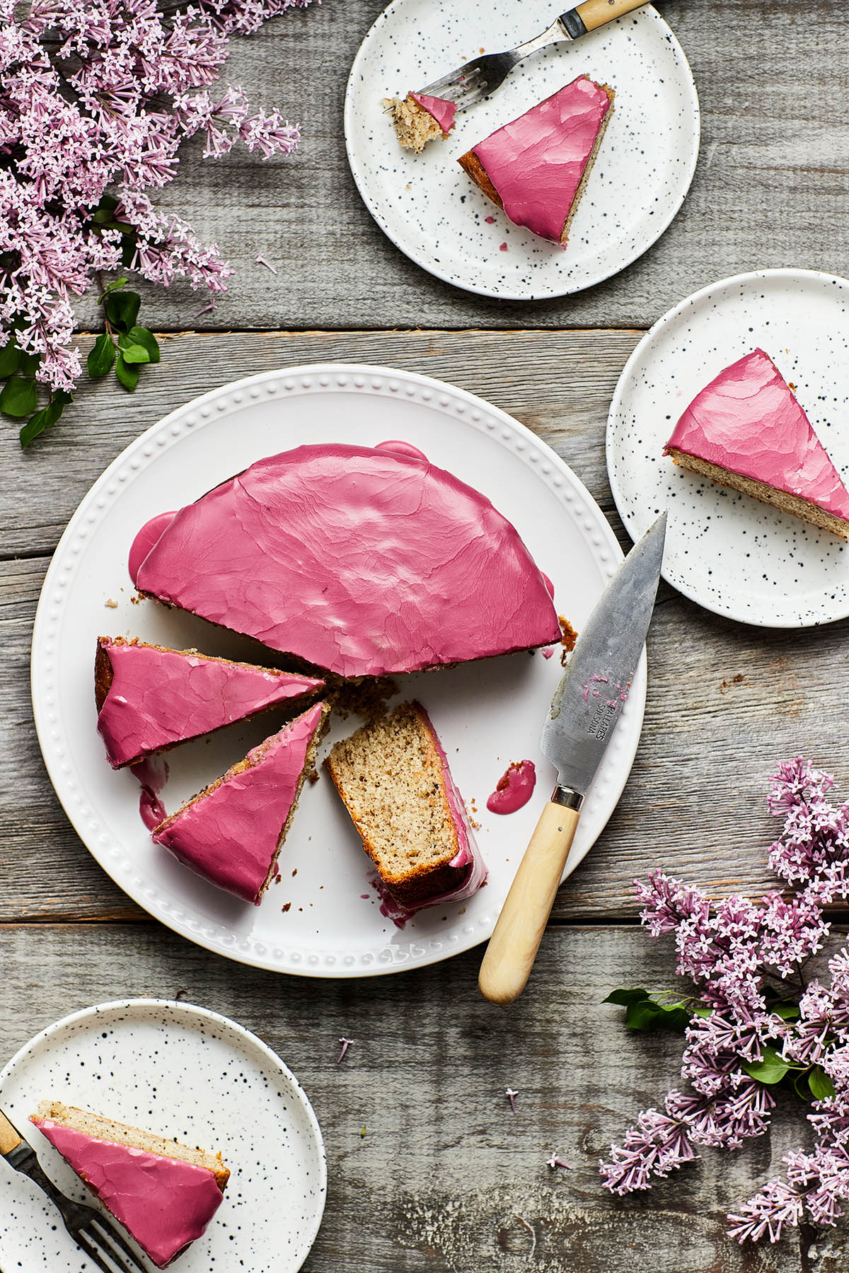 Overhead image of a sliced Earl Grey yogurt cake on a grey weathered table with slices of cake on small plates and fresh lilacs on the table around the cake.