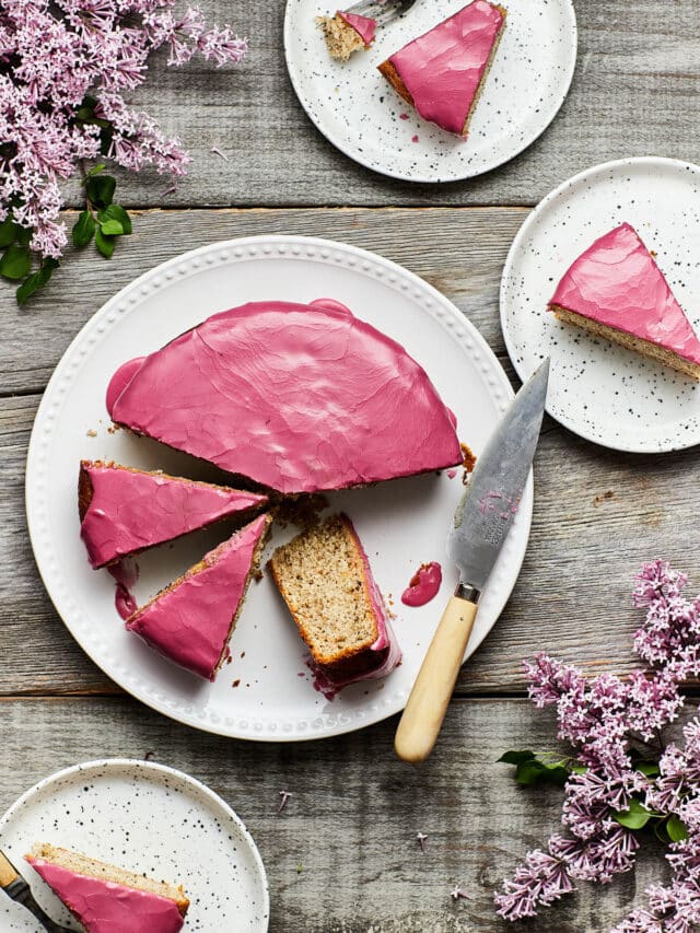 Overhead image of a sliced Earl Grey yogurt cake on a grey weathered table with slices of cake on small plates and fresh lilacs on the table around the cake.