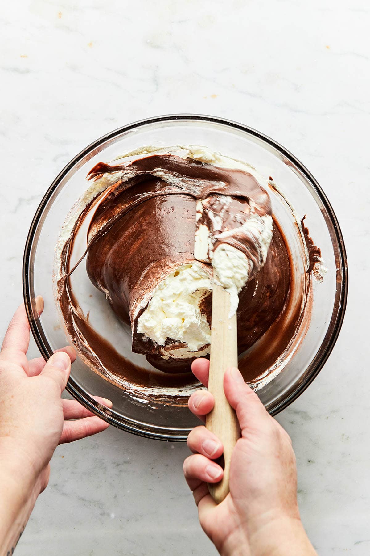 A hand using a rubber spatula to fold whipped cream into chocolate batter.