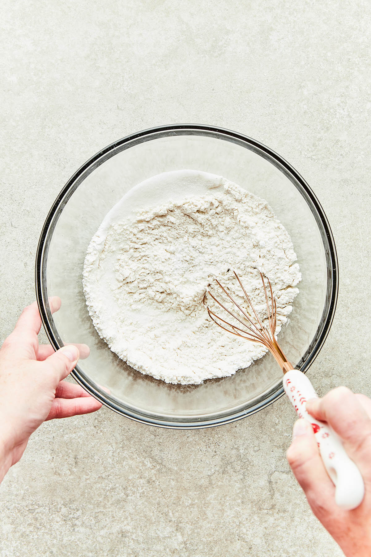 A hand using a whisk to mix dry ingredients in a large glass bowl.