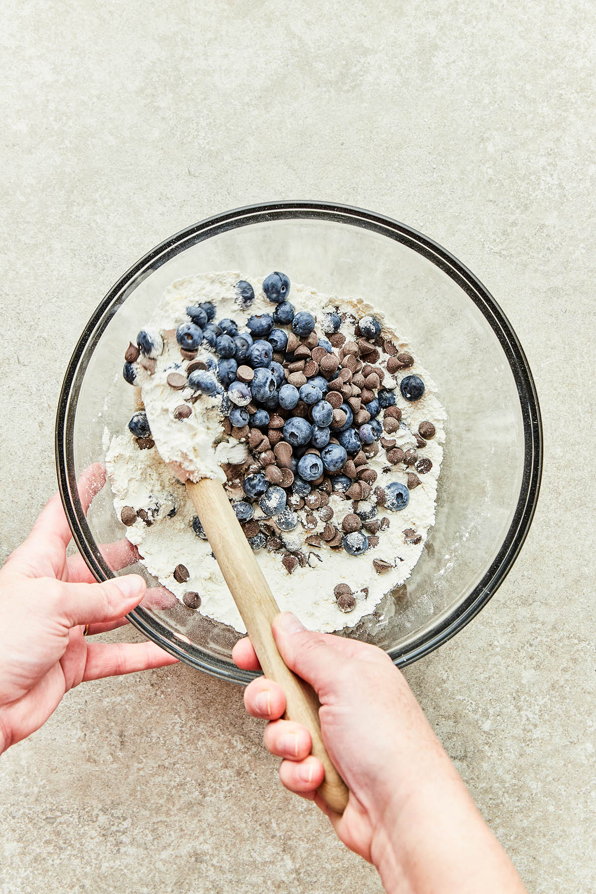 A hand using a rubber spatula to fold chocolate chips and blueberries into a bowl of dry ingredients.