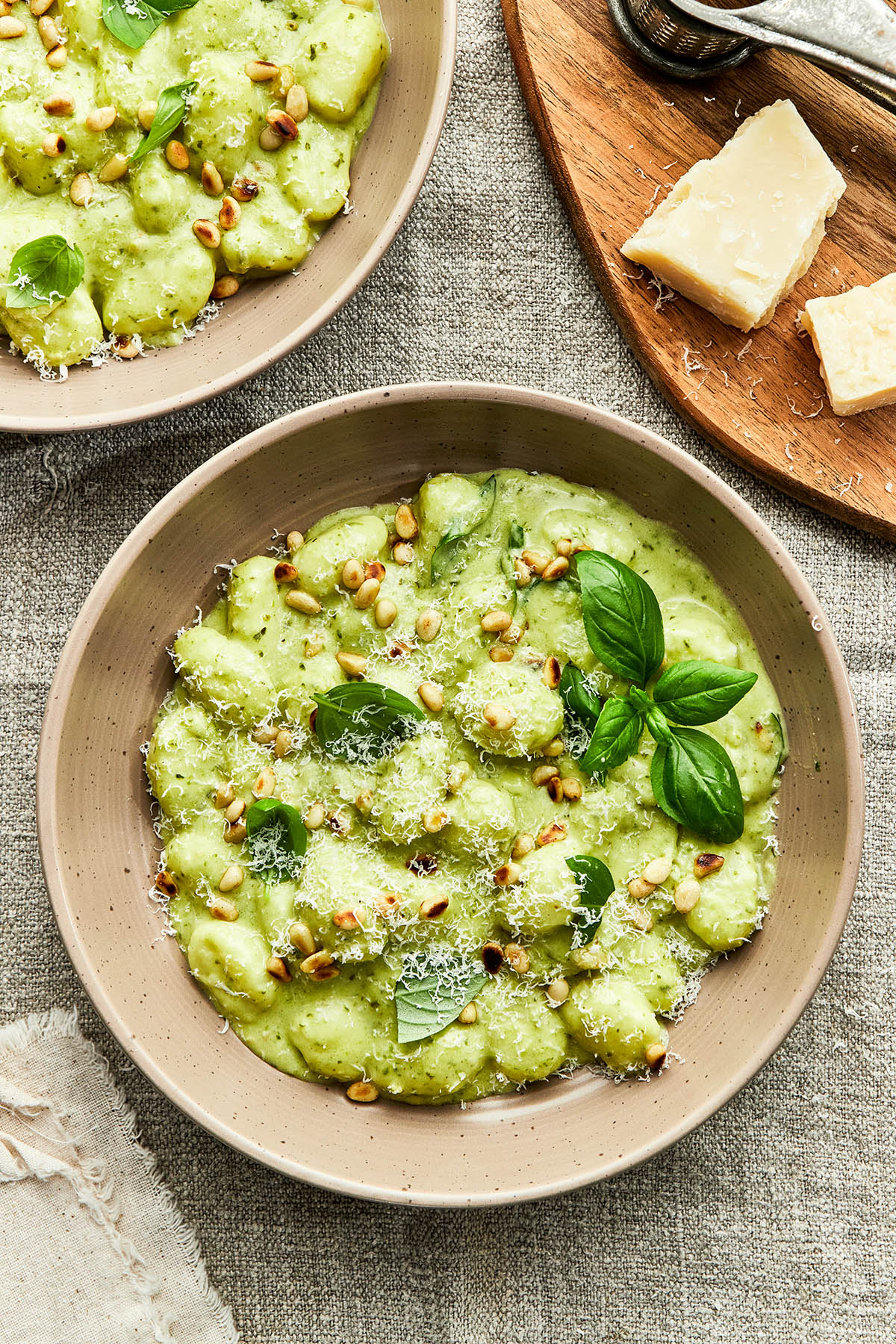A bowl of creamy pesto gnocchi on a linen tablecloth with another bowl of pasta and a wooden cutting board with chunks of Parmesan cheese nearby.