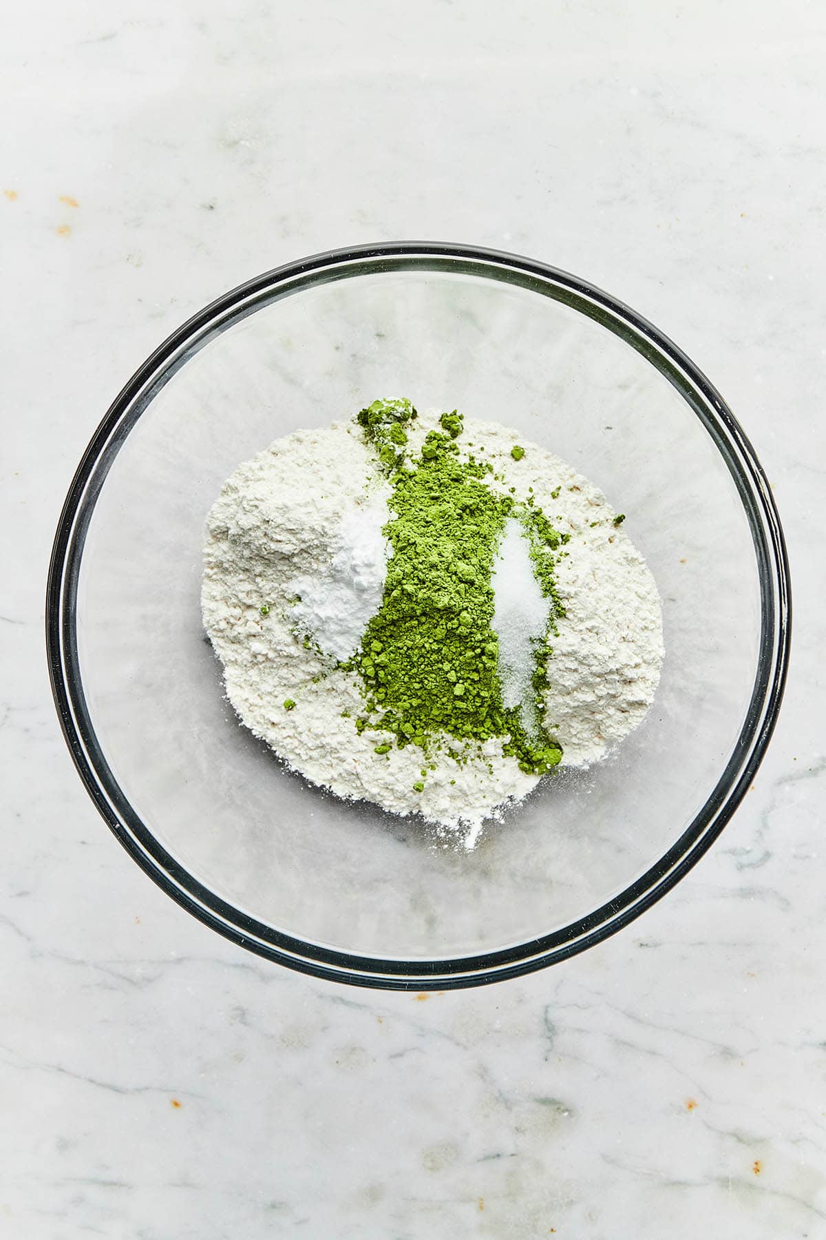 A bowl of unmixed flour, matcha powder, baking soda, and salt in a glass mixing bowl.