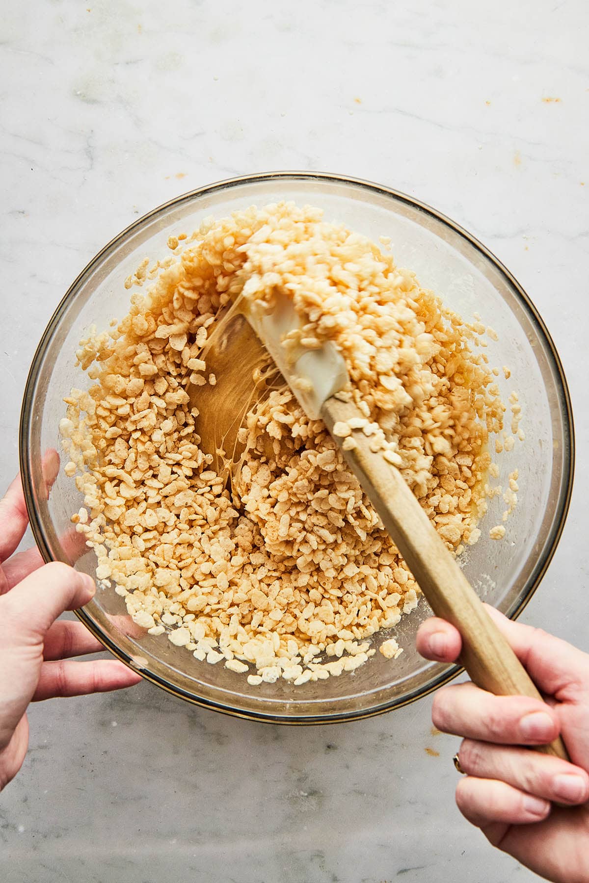 A hand stirring Rice Krispies ceral into a bowl of melted marshmalllow and peanut butter in a mixing bowl.