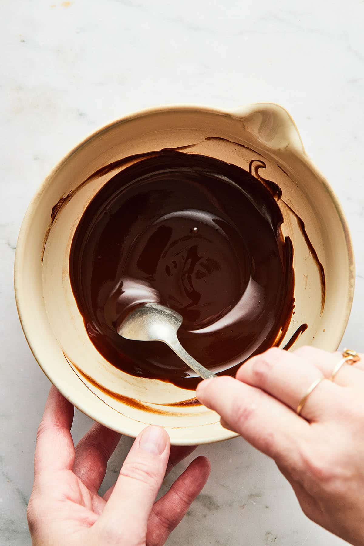 A hand using a spoon to stir chocolate glaze in a small mixing bowl.
