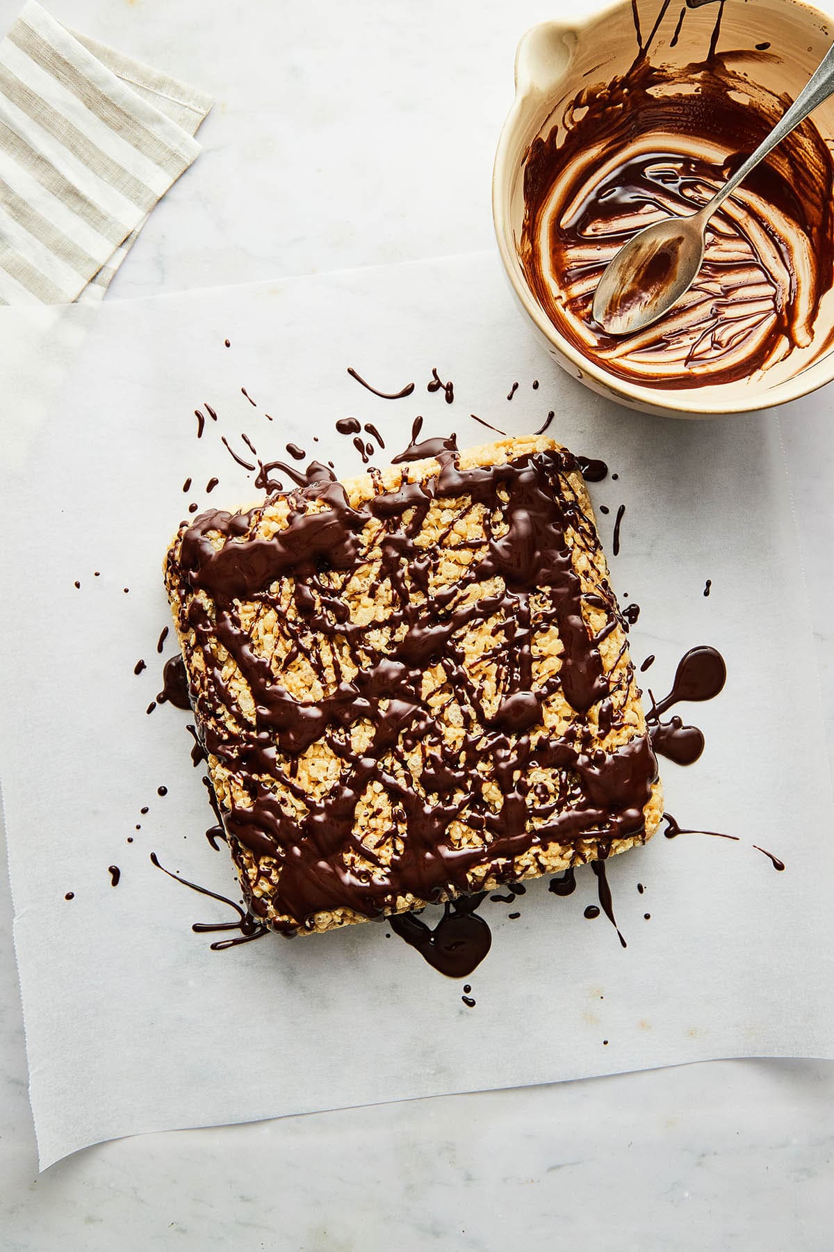 An unsliced square slab of unsliced marshmallow cereal bars drizzled generously with chocolate with a small empty bowl of glaze and a striped kitchen towel nearby.