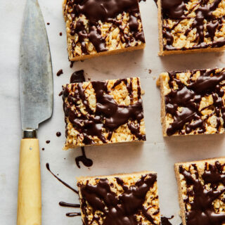 Six peanut butter Rice Krispie treats on a marble surface lined with white parchment paper. A knife is on the paper next to the squares.