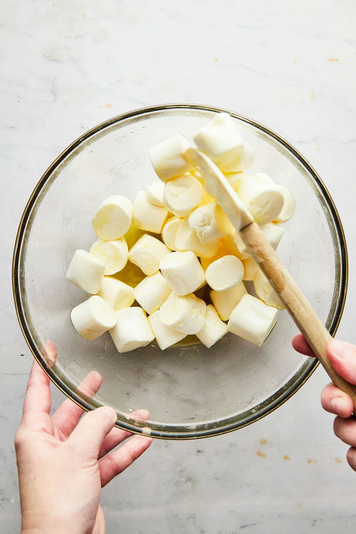 A hand stirring large marshmallows in melted butter, inside a glass mixing bowl, using a rubber spatula.