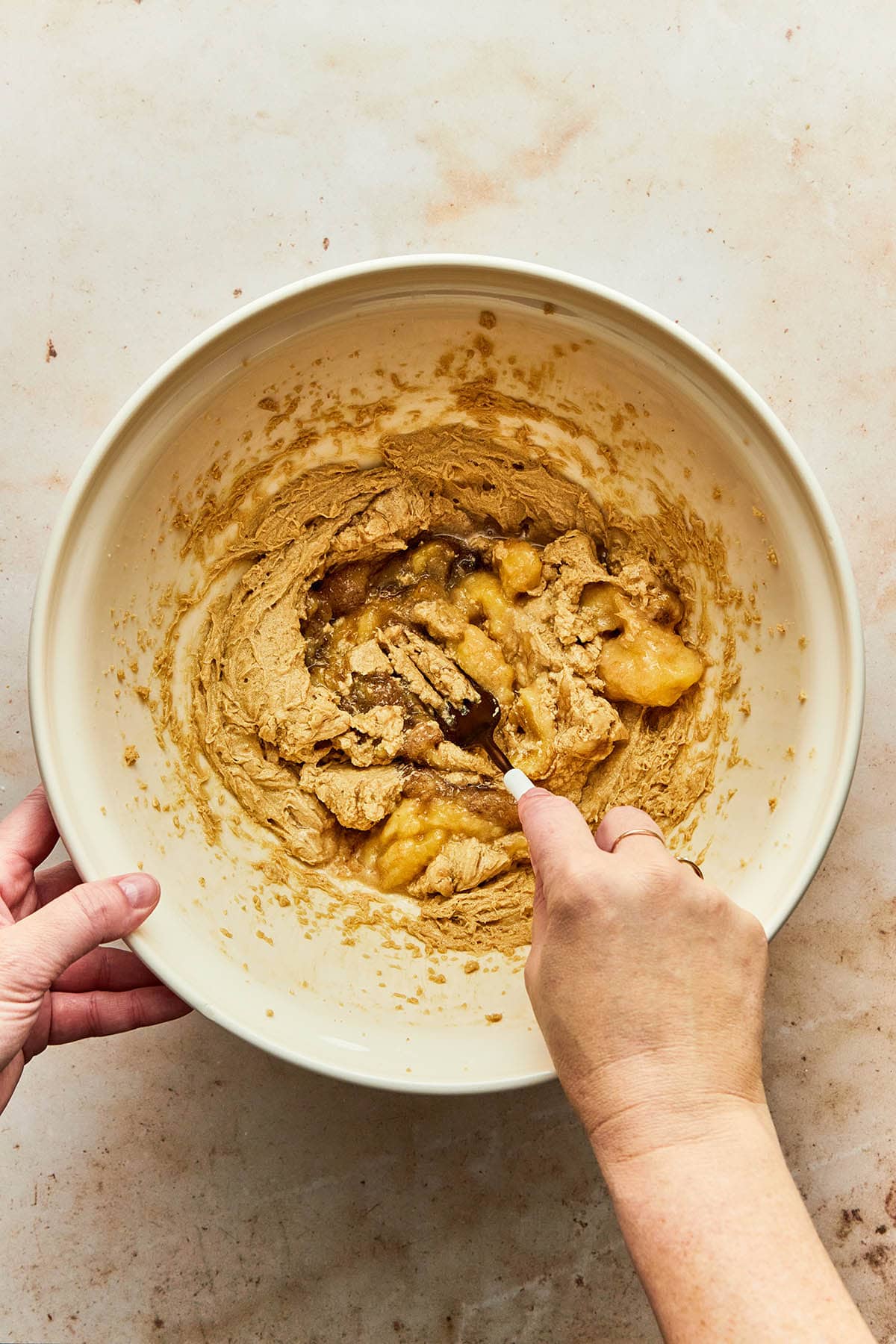A hand using a fork to mash overripe bananas into a bowl of wet muffin ingredients.