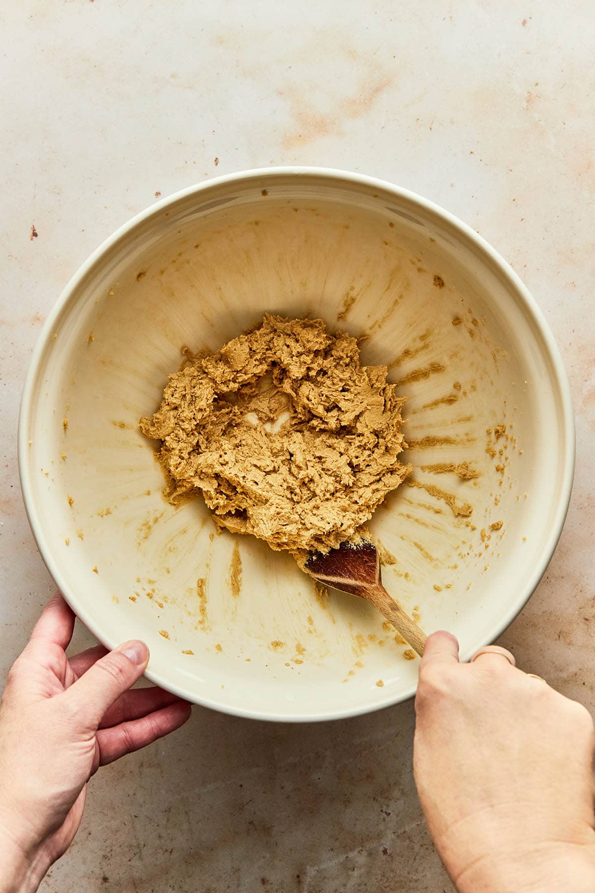 A hand using a wooden spoon to scrape down the sides of a mixing bowl.