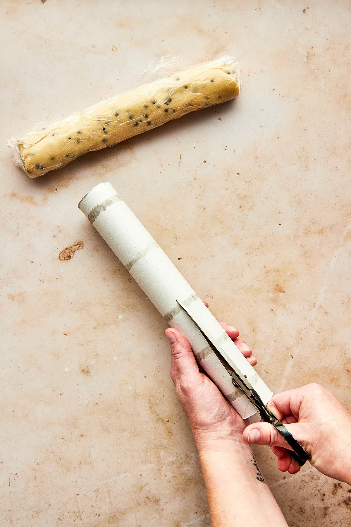 Hands using scissors to cut a cardboard paper towel tube down it's length. A wrapped log of cookie dough is nearby.