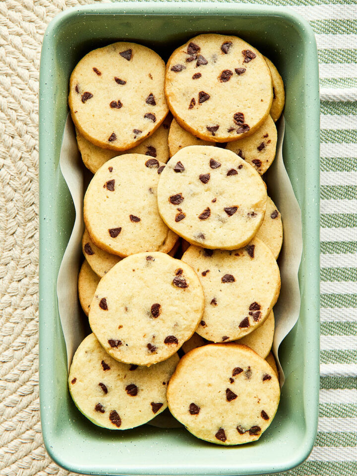 Chocolate chip sugar cookies in a mint green loaf tin lined with a white parchment paper circle on a green and white striped background with a kintted trivet covering part of the background.