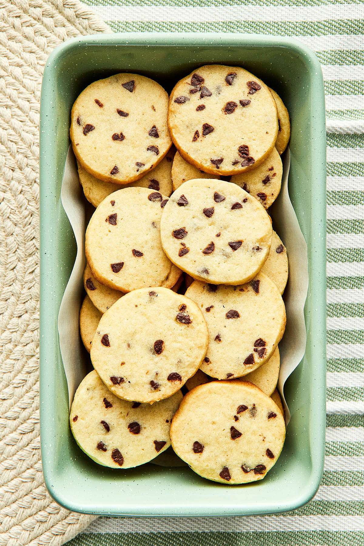 Chocolate chip sugar cookies in a mint green loaf tin lined with a white parchment paper circle on a green and white striped background with a knitted trivet covering part of the background.