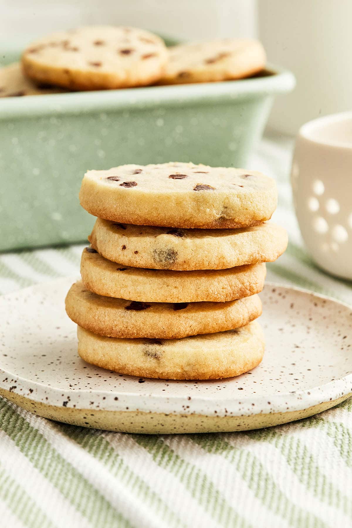 Front on image of a stack of chocolate chip sugar cookies on a small ceramic plate with a mint green tin of more cookies in the background.