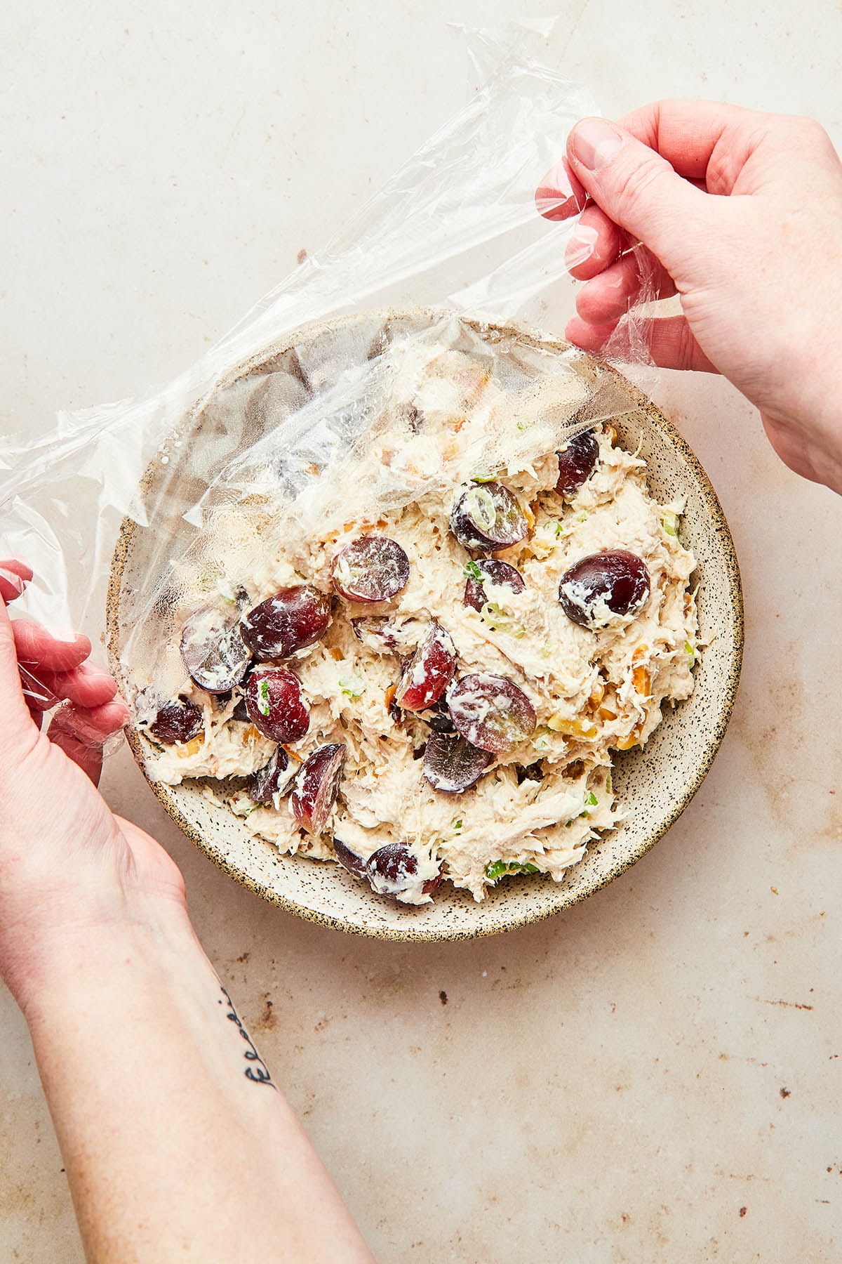 Hands removing plastic wrap from a bowl of easy chicken salad.