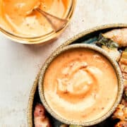 A jar of bang bang sauce next to a small platter of bacon wrapped shrimp with a dish of sauce on the plate.