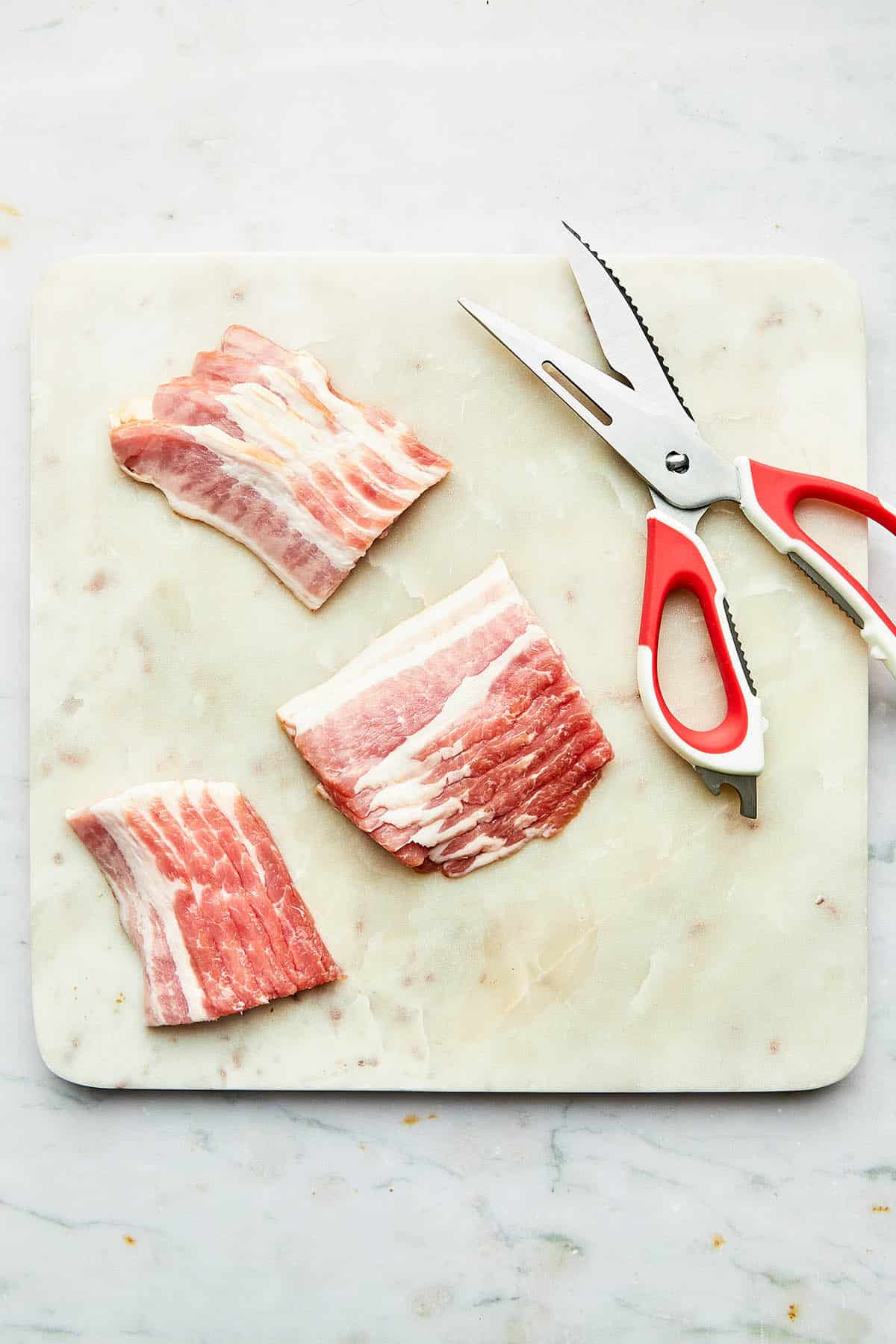 Bacon strips cut into thirds on a marble cutting board with a pair of kitchen shears nearby.
