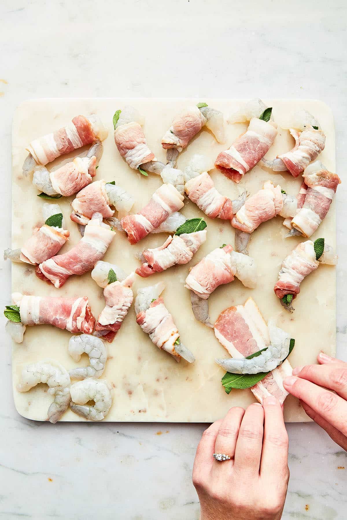 Hands about to roll a shrimp topped with a piece of fresh sage inside a small strip of bacon.