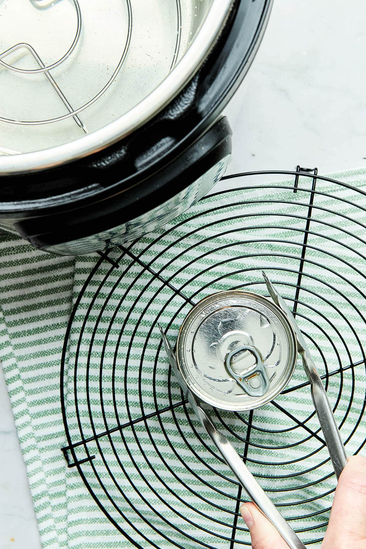 A hand using tongs to place a small can on a wire cooling rack with a green and white striped tea towel underneath.