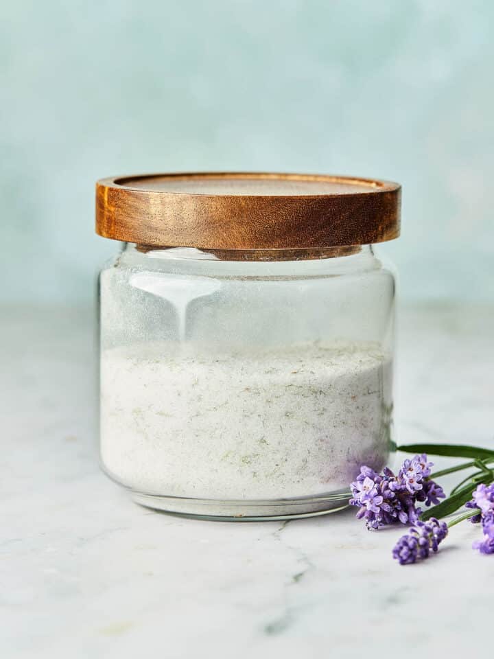 A glass jar of lavender sugar with a wood top and fresh lavender flowers on the table nearby.