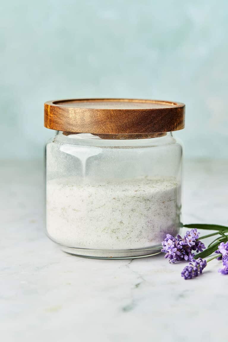A glass jar of lavender sugar with a wood top and fresh lavender flowers on the table nearby.