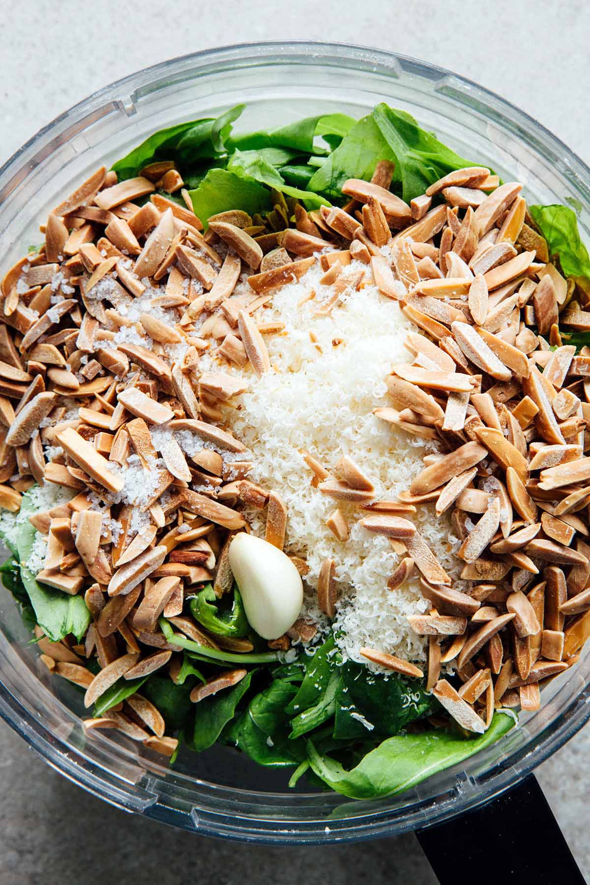The bowl of a food processor filled with fresh spinach and arugula, Parmesan cheese, toasted slivered almonds, flaky salt, and one clove of peeled garlic.