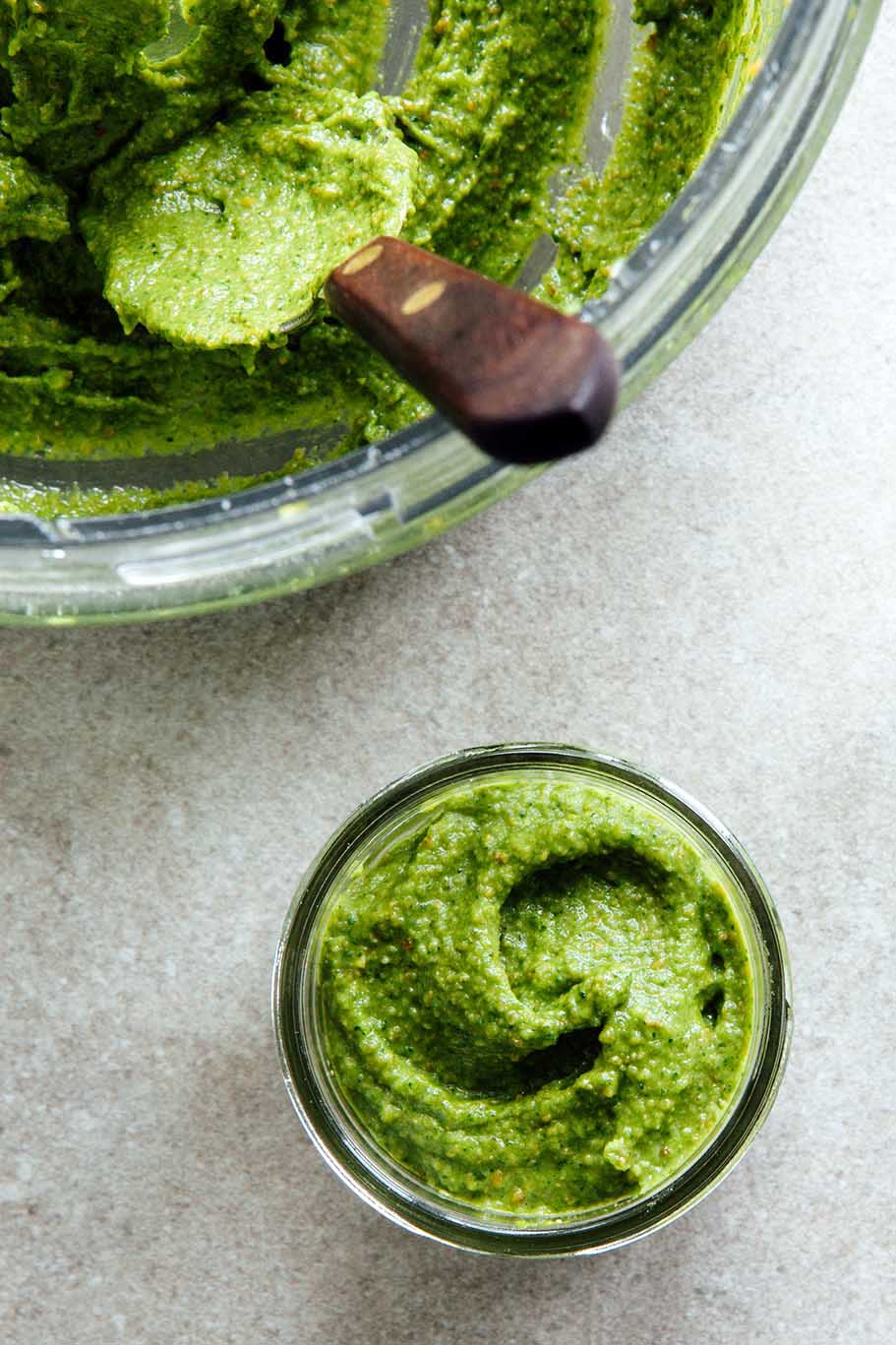 Small jars of pesto next to the bowl of a food processor with more pesto and a spoon inside of it.