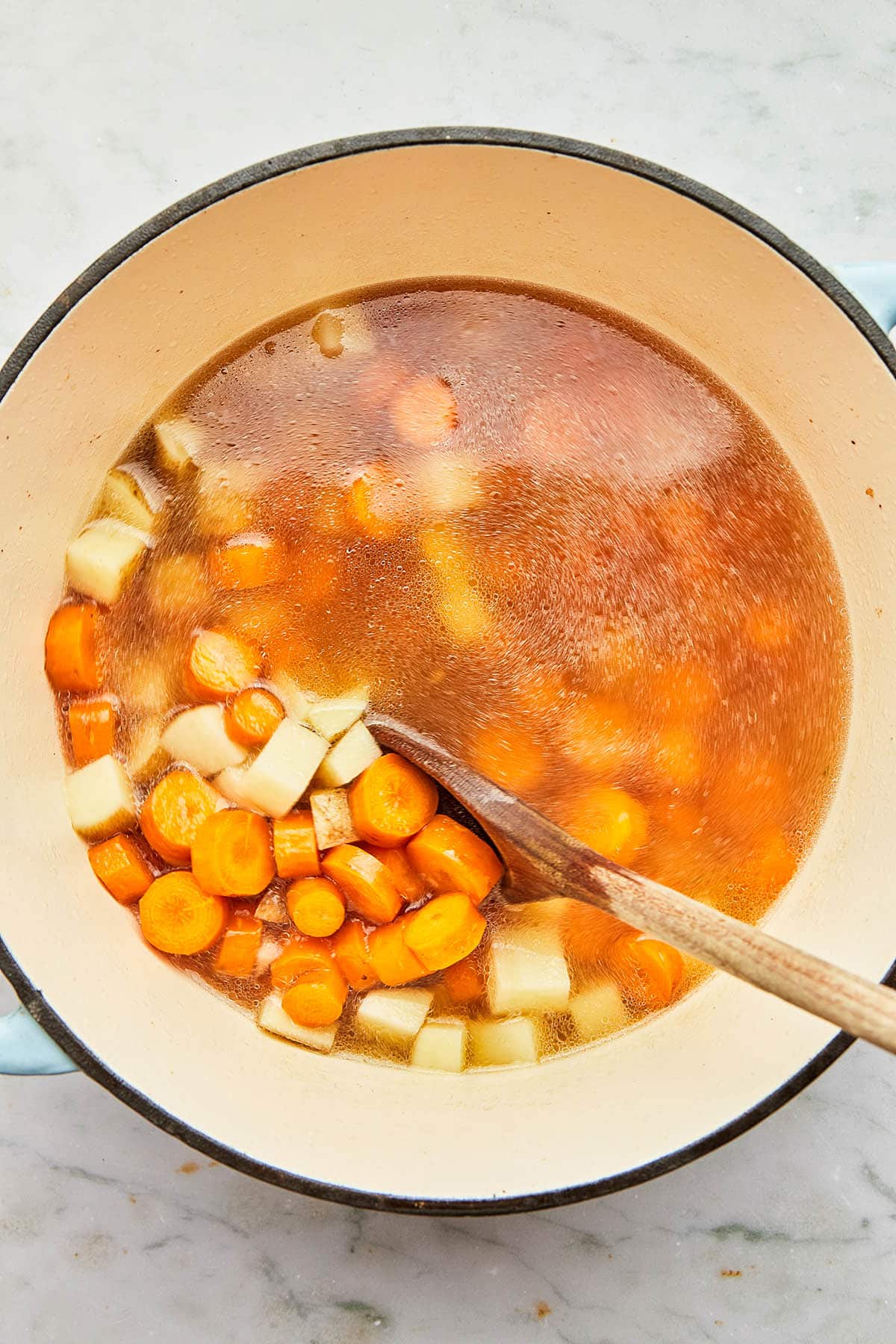 A wooden spoon stirring a pot of water with cooked carrots and potatoes inside.