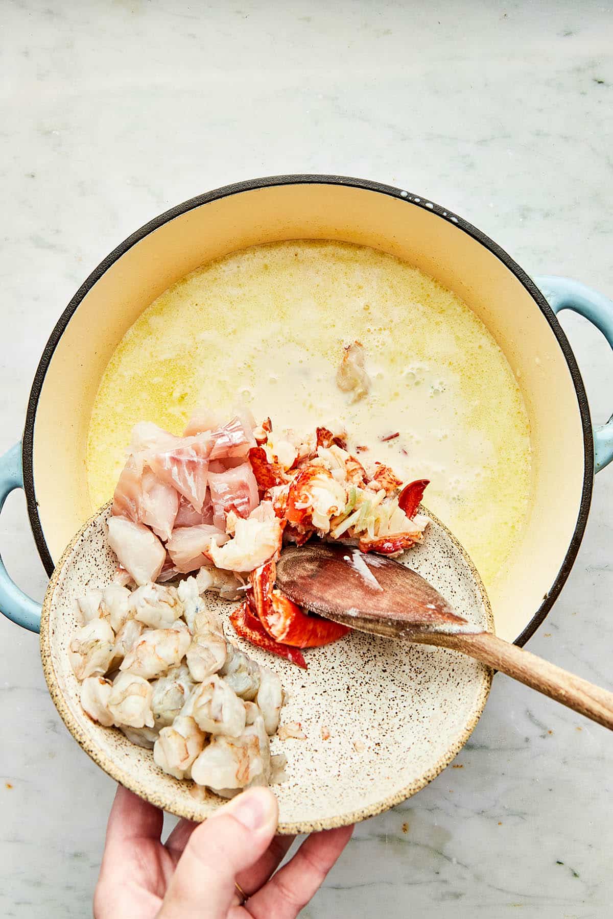 A hand using a wooden spoon to nudge seafood from a bowl into a pot of creamy soup.