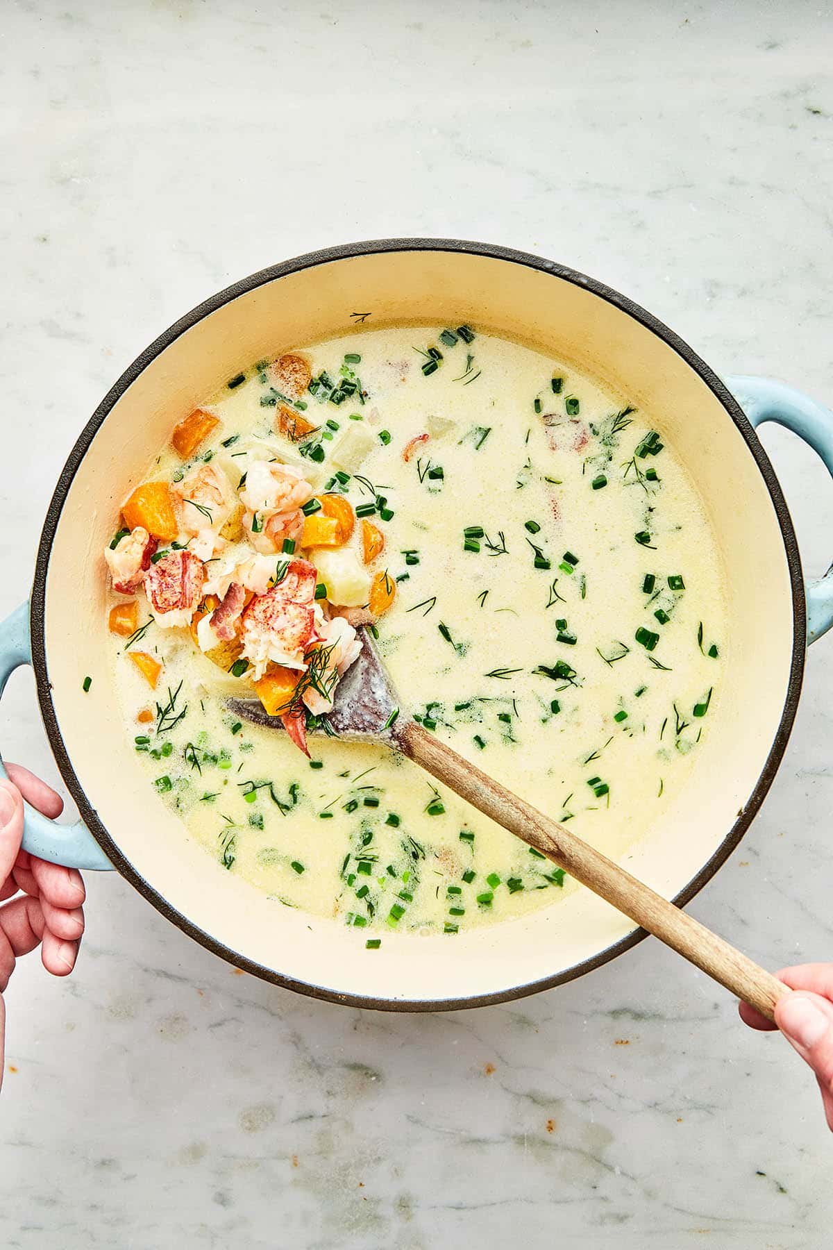 A hand using a wooden spoon to stir seafood chowder in a cast iron pot.