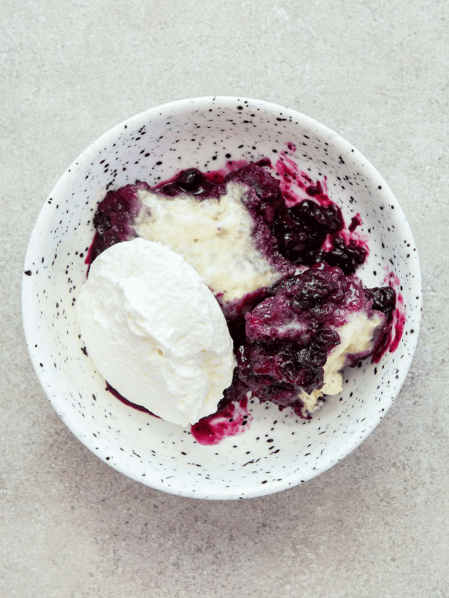A serving of Nova Scotia blueberry grunt topped with whipped cream in a white speckled bowl on a stone surface.