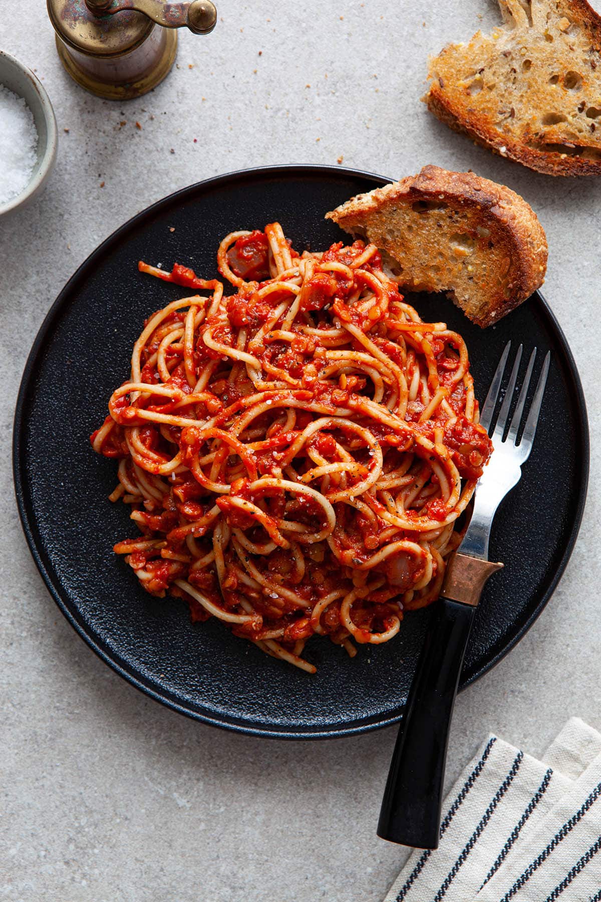 A plate of spaghetti tossed with lentil Bolognese sauce on a black plate with a black fork and a piece of toasted bread on the plate.
