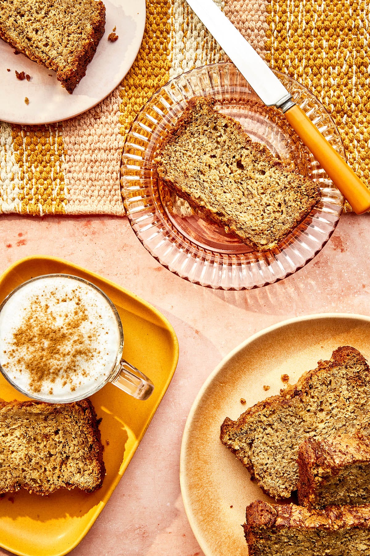 Slices of homemade quick bread on plates with a cappuccino dusted with cinnamon and a yellow-handled butter knife nearby.