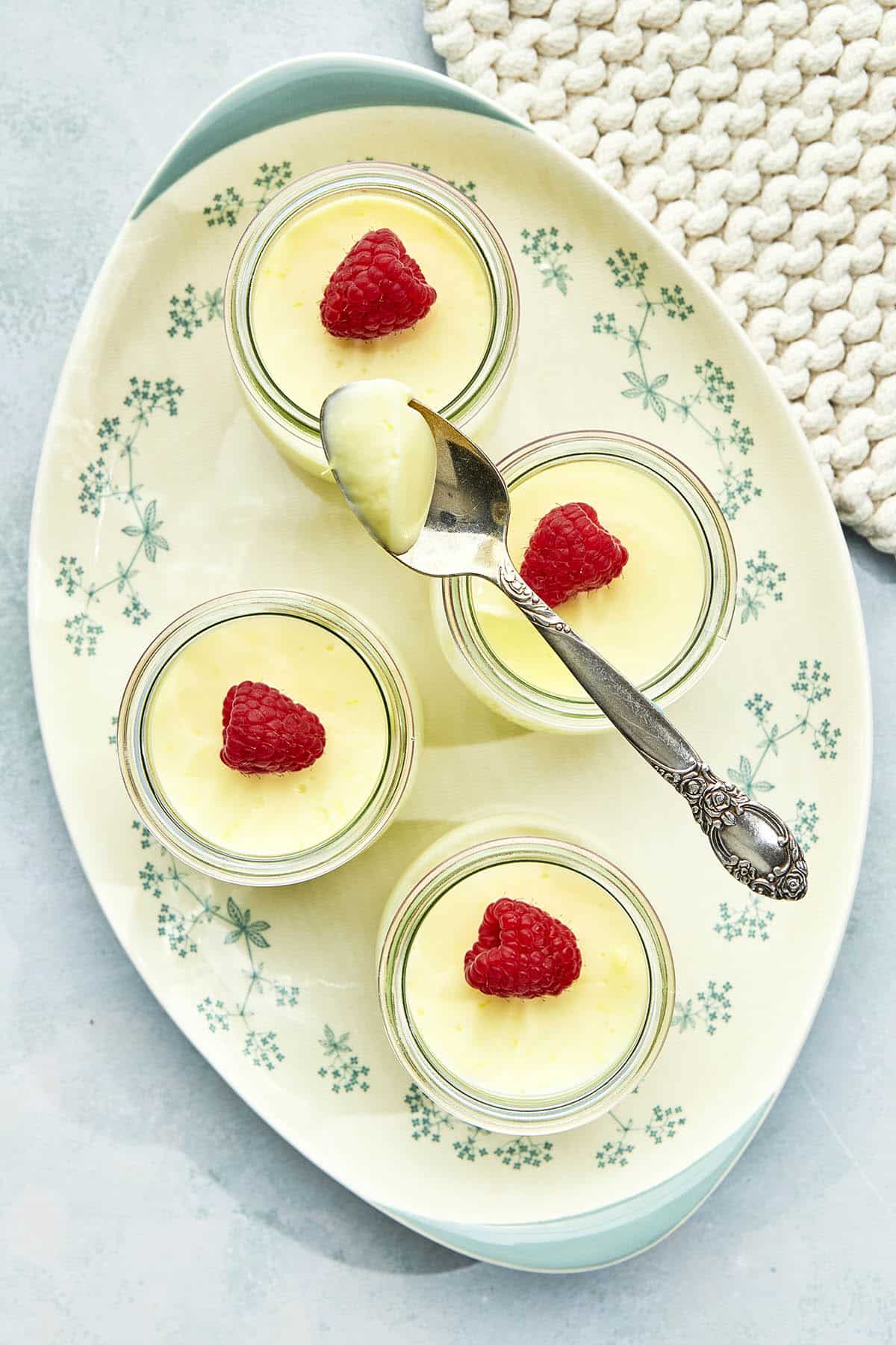 Four jars of lemon posset on an oval platter decorated with light blue flowers, with one spoon on top of one of the jars with some of the dessert spooned out.