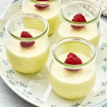 Four small jars of lemon posset, each pudding topped with a fresh raspberry, on a white oval platter decorated with light blue flowers.