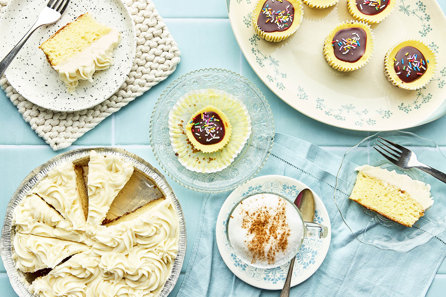 An aqua coloured tile background topped with around a vanilla cake with frosting in a metal tin, plates of singleserving desserts, and a small platter of mini cheesecakes.
