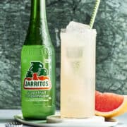 A Paloma Mocktail with a green and white striped straw and a bottle of grapefruit soda and a wedge of grapefruit nearby.