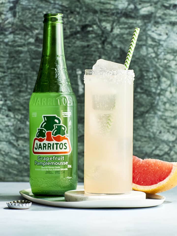 A Paloma Mocktail with a green and white striped straw and a bottle of grapefruit soda and a wedge of grapefruit nearby.