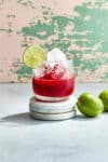 A raspberry margarita Mocktail sitting on three round marble coasters. The glass is filled with ice and has a salted rim and a lime wheel for garnish. The margarita itself is a vibrant rich red color and two limes sit nearby on the counter.