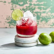A raspberry margarita Mocktail sitting on three round marble coasters. The glass is filled with ice and has a salted rim and a lime wheel for garnish. The margarita itself is a vibrant rich red color and two limes sit nearby on the counter.